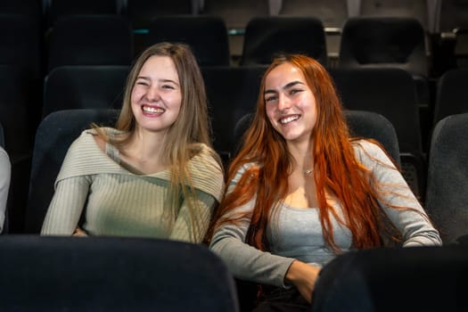 Two happy young women are sitting in comfortable cinema seats, enjoying a movie and smiling