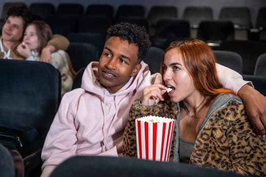 Young multi ethnic couple enjoying a movie and sharing popcorn in a cinema, surrounded by other people