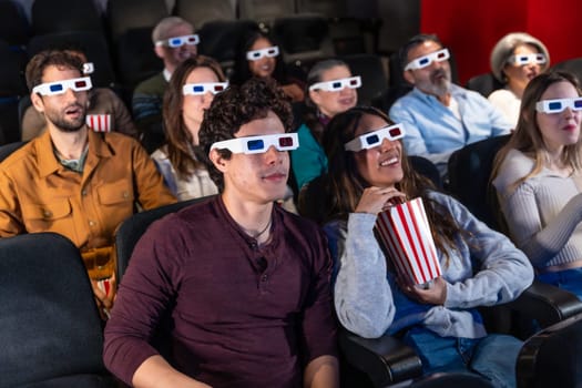 People of different ages wearing 3d glasses are watching a movie in a cinema, eating popcorn and enjoying the show