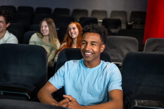 Young man smiles while watching a movie in a cinema, surrounded by his friends, enjoying a shared entertainment experience