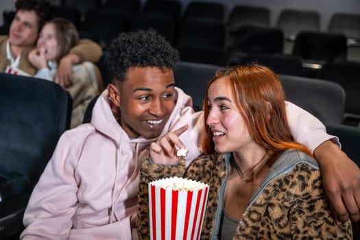 Happy diverse couple enjoying film and sharing popcorn bucket in movie theater, whispering during the projection