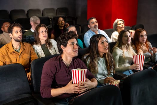 People of different ages and ethnicities are watching a film in a movie theater, holding popcorn buckets and enjoying the show
