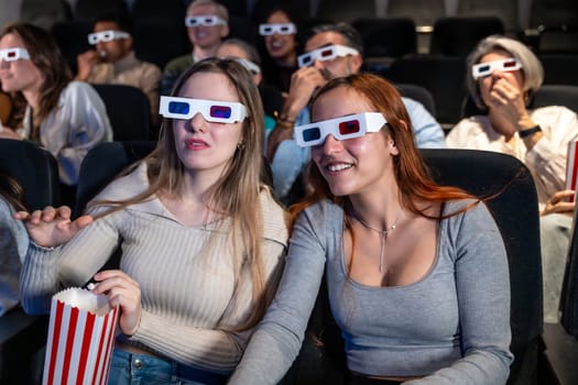 Two young women wearing 3d glasses are watching a movie and eating popcorn in a crowded movie theater
