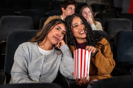 Two friends enjoying a movie at the cinema, one is eating popcorn while the other looks bored