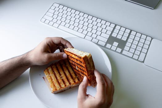 Male hands holding grilled sandwich near computer keyboard on desk.