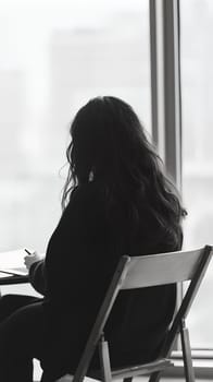 Black and white photo of a woman sitting by a window, deep in thought, with a serene backdrop of the city.
