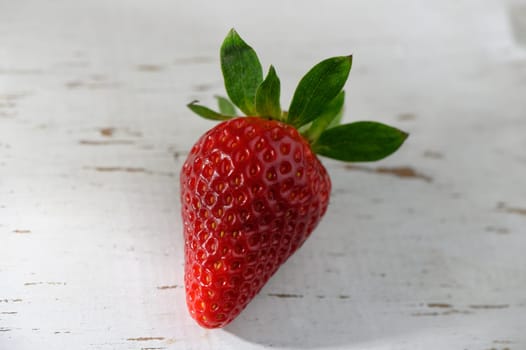 A vibrant red strawberry rests on a weathered wooden table, illuminated by gentle sunlight. The fruit showcases its shiny surface and green leaves, symbolizing freshness and summer bounty.