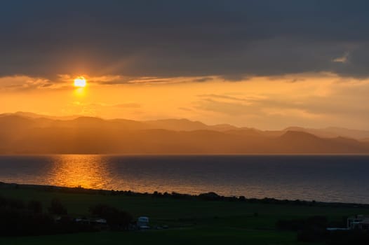 As the sun dips below the horizon, rich golden hues paint the sky, reflecting on calm waters. Distant mountains add depth to the serene landscape, creating a moment of pure tranquility.
