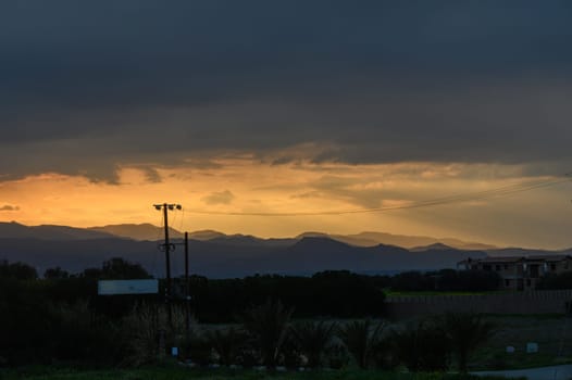 A breathtaking view as the sun sets behind the mountains, casting a warm glow against dark clouds. Power lines crisscross the landscape while shadows of trees emerge in the foreground.