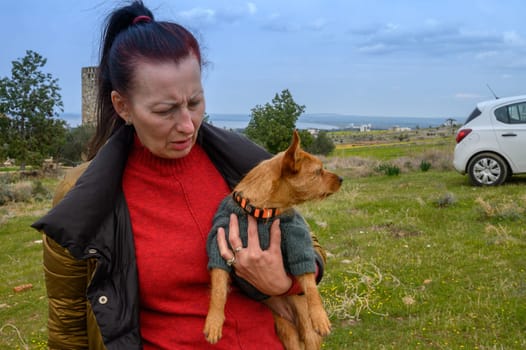 In a tranquil outdoor setting, a woman holds a small dog close, gazing thoughtfully into the distance. Green grass and distant hills create a serene backdrop as clouds drift above.