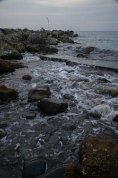 As daylight fades, the gentle waves roll over smooth stones along a rocky shoreline, with soft ripples reflecting the blue-gray hues of the twilight sky, offering tranquility.