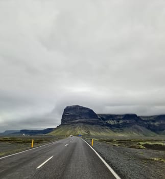 Asphalt road leading towards a large mountain in Iceland, under a cloudy sky, creating a dramatic and scenic view