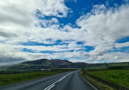 Asphalt road crossing green meadows with mountains and a cloudy sky in the background, typical Icelandic landscape