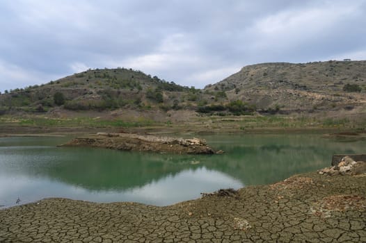 A once-thriving lake now shows signs of drought as muted green waters reflect the cloudy sky. The cracked earth reveals the harsh impact of dry conditions on this natural landscape.