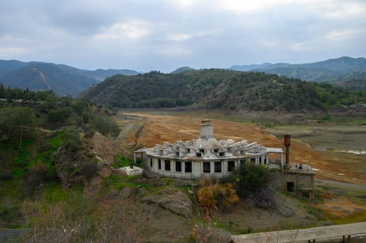 Once a bustling site, the decaying building stands against a striking landscape of parched earth and rolling hills, under a moody sky. Nature begins to reclaim its space.