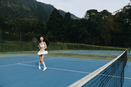 Happy young woman in white tennis attire enjoying a game on an outdoor tennis court, with lush green mountains in the background, embodying active lifestyle and sports enthusiasm
