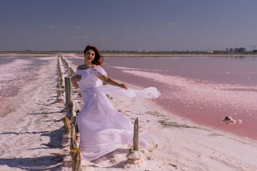 A woman in a white dress stands on a wooden walkway over a pink salt lake, her dress billowing in the wind.