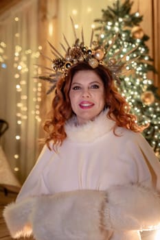 A woman with red hair and a white fur coat poses in front of a Christmas tree, wearing a golden crown of branches and ornaments.