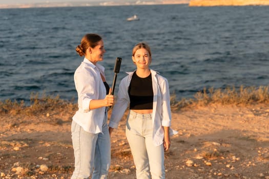 Two women, one holding a selfie stick, pose for a photo by the water. The ocean is behind them, with a small boat in the distance.