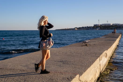 A blonde woman walks along a concrete pier overlooking the ocean. The water is blue and calm, with a few boats in the distance. The sky is clear and blue.
