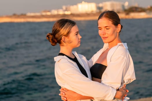Two women, one in a white shirt and black tank top, the other in a white shirt, embrace by a calm ocean with a cityscape in the background.