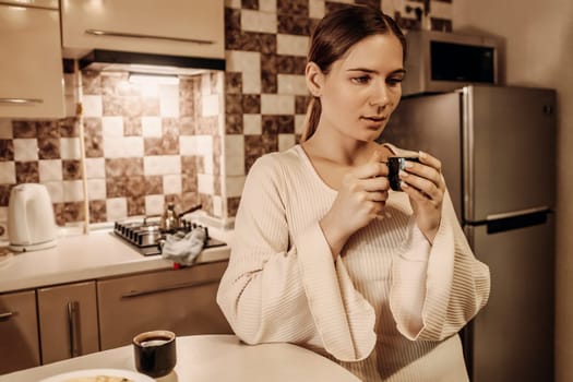 Woman in a cream robe examines a small dark object in her hands within a domestic kitchen setting. A cup and plate sit on the table nearby.