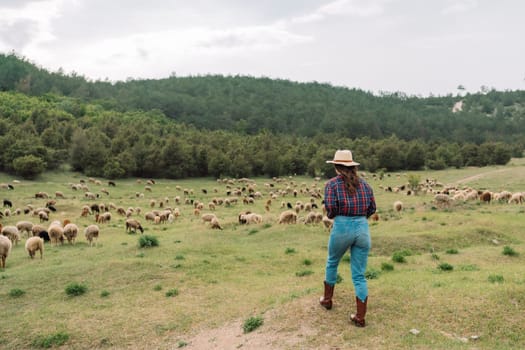A woman wearing a hat stands in a field of sheep. The sheep are scattered throughout the field, with some closer to the woman and others further away. Concept of peacefulness and tranquility