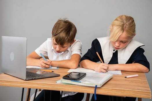A boy and a girl sit at a desk, both writing in notebooks. A laptop and a phone are on the desk. They are likely doing homework or studying.