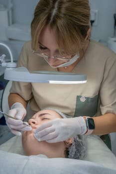 A beautician, wearing gloves and a mask, meticulously cleans a client's face using a small tool under a bright lamp. Facial treatment in progress.