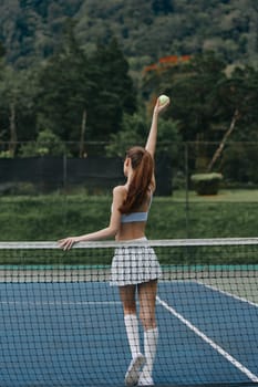 Energetic young female tennis player in a sporty outfit serving a ball on a sunny outdoor court, surrounded by lush greenery, portraying focus and athleticism
