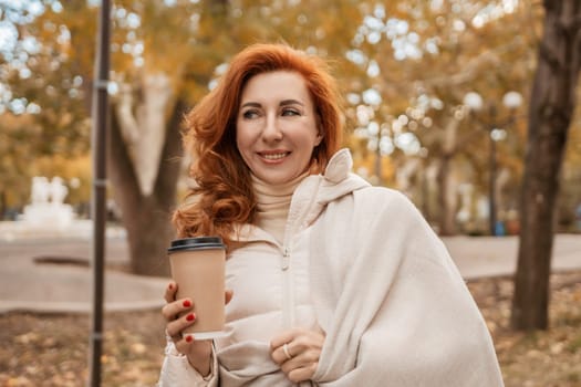 Redhead woman smiles, wrapped in a cream-colored shawl, holding a coffee cup outdoors in an autumnal park. Leaves are golden.