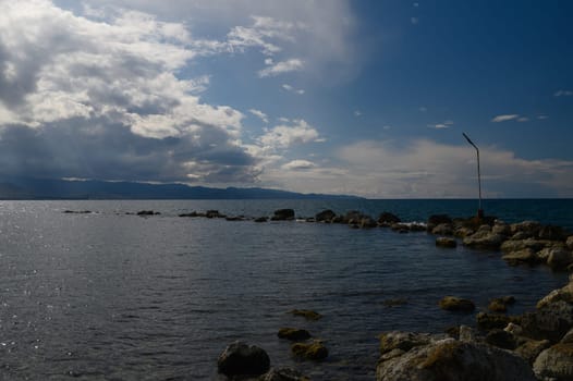 At the edge of a tranquil sea, smooth rocks emerge from the water under a vast sky filled with swirling clouds. The distant mountains create a stunning backdrop, enhancing the serene atmosphere.