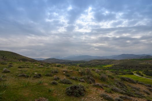 A tranquil landscape unfolds with rolling hills bathed in soft twilight. Lush greenery contrasts against an expansive sky filled with dramatic clouds, evoking a serene atmosphere of nature's splendor.