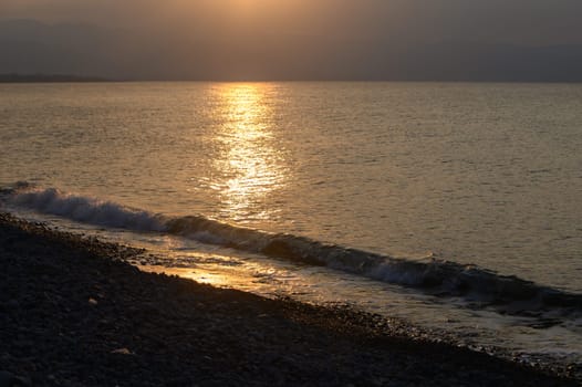 Golden sunlight glistens on gentle waves rolling onto a rocky beach, creating a peaceful ambiance as dusk approaches. The horizon blends colors beautifully in the evening sky.