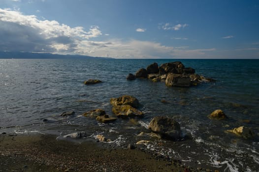 Waves rhythmically embrace the rocky shore, reflecting sunlight on the water's surface. Majestic clouds drift overhead, creating a peaceful and refreshing atmosphere by the ocean.