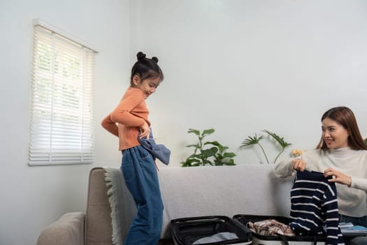 With smiles and laughter, the young girl eagerly stands on the couch to assist her mother in organizing their suitcase for an upcoming vacation.