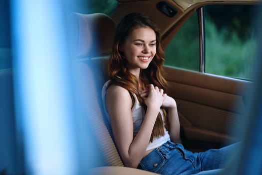 A joyful young woman in a white tank top and blue jeans sits in a car, exuding happiness and relaxation on a sunny day, surrounded by nature, embodying freedom.