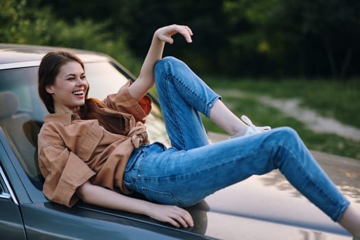 A young woman exudes happiness as she lounges on a vintage car, dressed in casual denim jeans and a trendy brown shirt, set against a vibrant green background, ideal for summer themes.