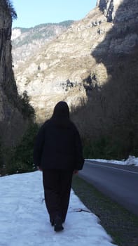 Person in black jacket walks away from camera on snowy path beside road; steep, rocky mountains loom in background.