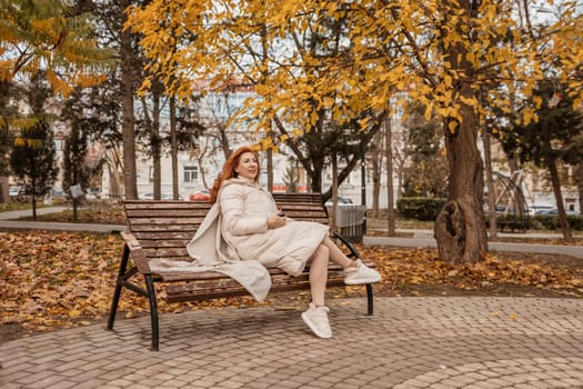Redhead woman sits on a park bench beneath golden autumn leaves. She wears a cream-colored sweater dress and white sneakers. Fallen leaves cover the ground.