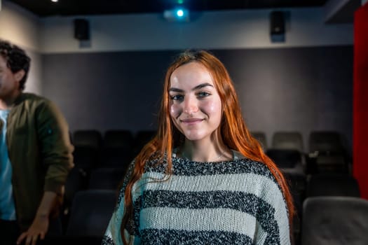 Redhead young woman smiling during movie session in a cinema theater, audience in the background