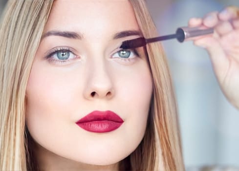 Woman applying mascara with confidence, defined brows and red lipstick. Blonde beauty focused on her eye makeup, cosmetics look