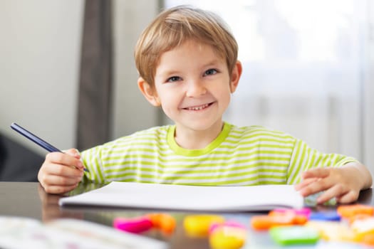 Young boy in a green striped shirt sitting at a table, smiling while holding a pencil and drawing on a white sheet of paper. Colorful educational toys are scattered around