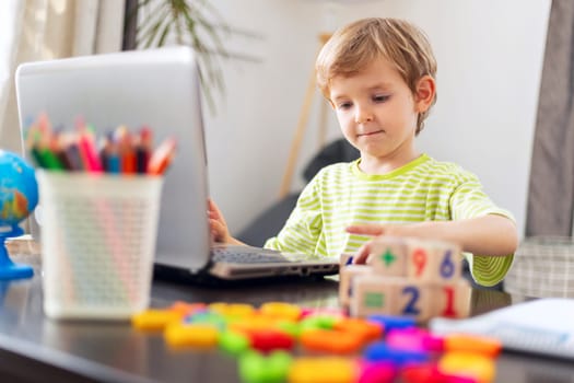 Young boy in a striped green shirt using a laptop at a desk with colorful educational toys, pencils, and a small globe. The child is engaged in remote learning in a home setting