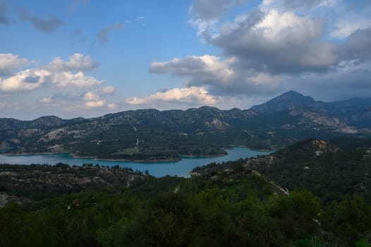 Lush green hills cradle a tranquil turquoise lake, while dramatic clouds drift above mountains at dusk, creating a breathtaking natural panorama.