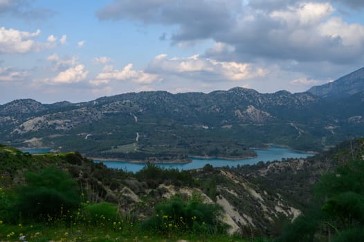 Rolling mountains cradle a shimmering turquoise lake as clouds drift across the sky at sunset. Wildflowers bloom along the slopes, enhancing the tranquil beauty of nature's masterpiece.