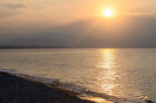 As the sun dips below the horizon, soft golden rays reflect off calm waters, illuminating a pebble-strewn shore. A gentle breeze stirs the peaceful atmosphere at twilight.