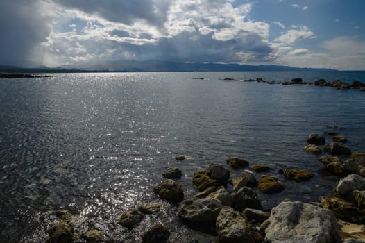 Waves gently lap against a rugged shoreline scattered with rocks and moss. A dramatic sky filled with clouds casts shifting shadows, creating a serene atmosphere at sunset.