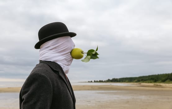 A man with a face covered by a cloth, wearing a hat and a coat, is holding an apple in his mouth without using his hands on the seashore.