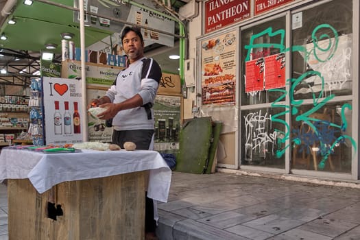 Crete, Greece - April 5, 2025: A vendor presents a kitchen tool at a busy market stall with colorful vegetables and promotional posters in the background.